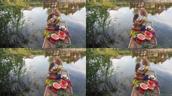 妈妈和小女儿穿着牛仔短裤和背带，头上戴着红色的蝴蝶，坐在河边的钓鱼平台上，拥抱亲吻