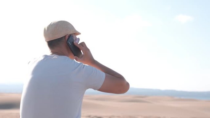 一个人独自坐在沙漠沙丘上，用智能手机打电话。复制空间