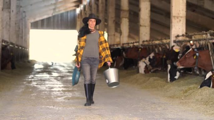 4k视频宽镜头，女农场工人拿着水桶和铁锹在农场牛棚里行走