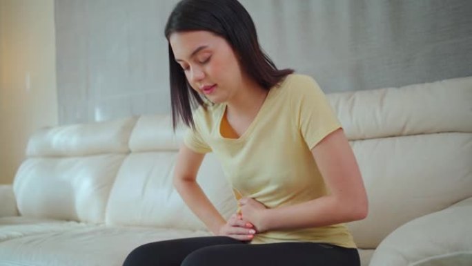 亚洲迷人的女孩痛苦的脸，紧握的腹部感觉患有严重的腹痛。紧张的年轻女子坐在家里的沙发上，肚子疼或月经。