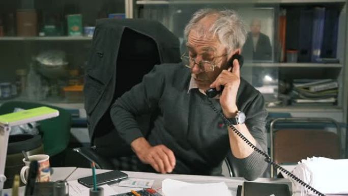 一位老人坐在办公桌前，用固定电话打了电话。