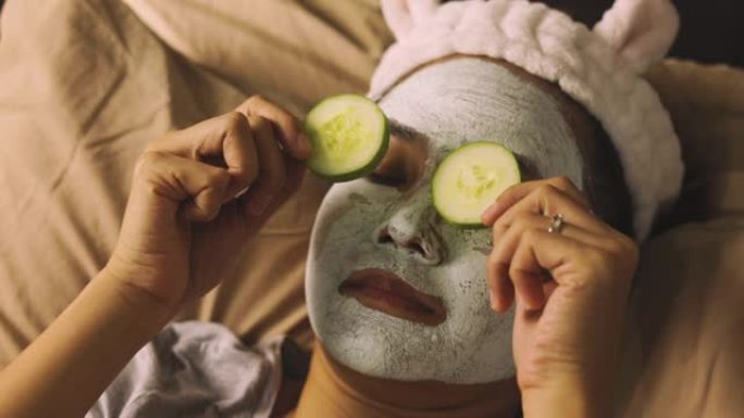中等年龄亚洲女性的面部聚焦真实动作，她用泥遮住脸去除眼睛上的黄瓜切片，滋润床上皮肤，放松温暖的光线。