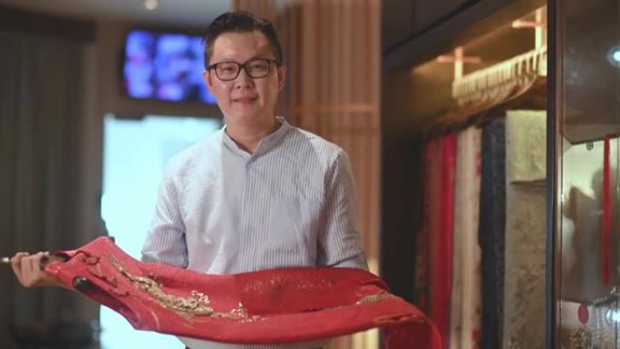 自信的男性中国传统服装时装设计师拿着传统服装旗袍看着相机微笑着在他的设计工作室精品店