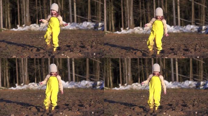 女孩走进泥泞。在地面的水坑里跺脚，玩有趣的游戏。快乐的孩子在春天在乡村森林里玩耍。黄色防水连身衣，橡