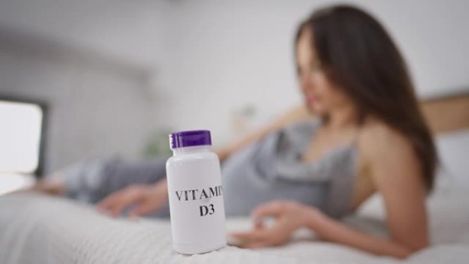 维生素D3瓶躺在床上，背景是模糊的孕妇看书。深色皮肤的高加索年轻人在室内享受业余爱好，前面有药丸。保