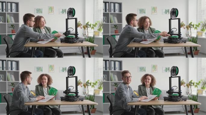 现代女性和男性的肖像在家里使用3D打印机，手里拿着3D模型，坐在房间的桌子上，微笑着看着相机