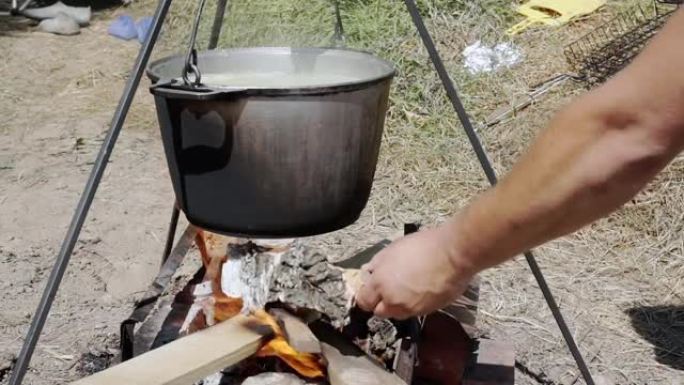 一名男子将柴火放在火上，在背景中: 一锅着火，做饭