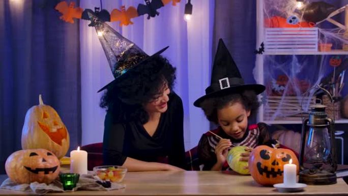 穿着节日服装和巫婆帽子的小女孩和女人非洲裔美国人会画南瓜。妈妈和女儿坐在万圣节之夜装饰的房间的桌子旁