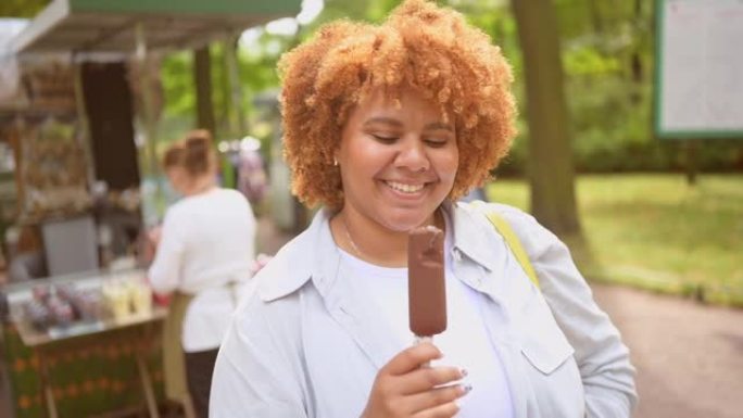 微笑的年轻自然美丽的又大的非洲裔美国妇女在公园吃冰淇淋巧克力冰棍的生活方式肖像。多样性和身体积极的概