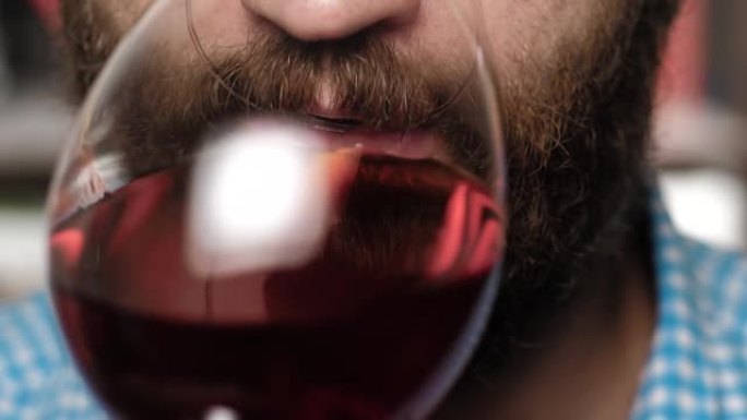 大胡子的人正在喝酒。男性的手将玻璃杯带到他的嘴里，喝红酒。特写