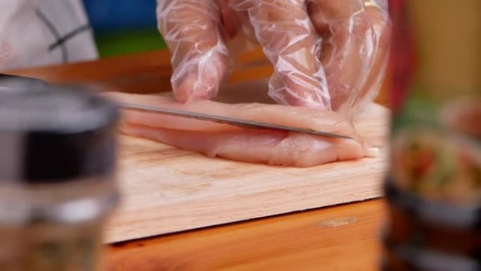 戴着手套的女人正在木制砧板上切鸡肉，准备好吃的食物。自制烹饪概念。