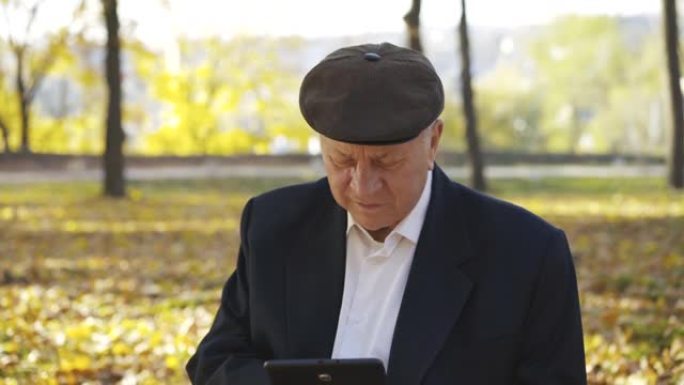 戴帽子的高级男子使用平板电脑，并在阳光明媚的秋天背景下讲述