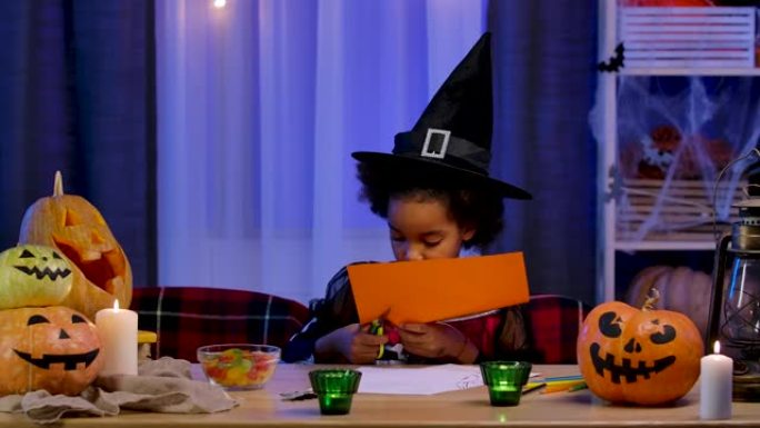 穿着节日服装和巫婆帽子的非洲裔美国小女孩用橙色纸切南瓜。孩子坐在万圣节之夜装饰的房间的桌子旁。慢动作