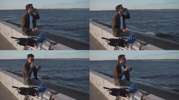 骑自行车的人坐在海边的水泥栅栏上，喝一次性咖啡，骑自行车时吃三明治。男子在湖边骑自行车后吃了午饭。下