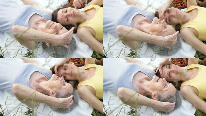 美丽的老祖母，白发和皱纹的脸正躺在白色地毯上，她的成年女儿在户外野餐，母亲节，退休快乐