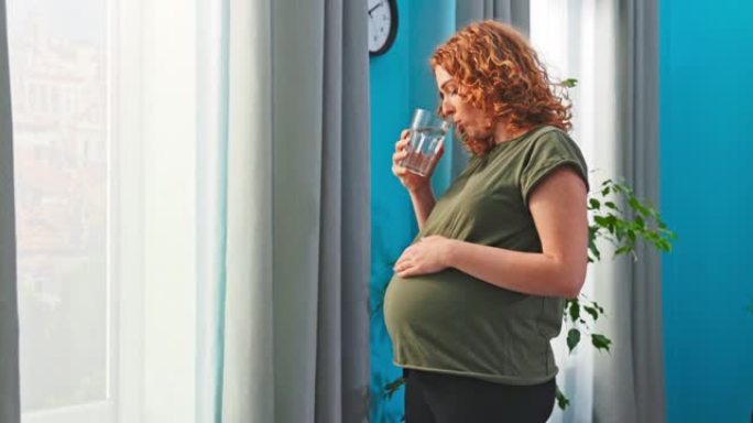 年轻的准妈妈站在客厅的窗户旁边，手里拿着一杯水。女孩喝水，注意补水，怀孕期间饮食良好，补充矿物质，中