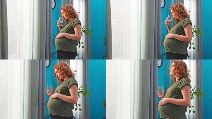 年轻的准妈妈站在客厅的窗户旁边，手里拿着一杯水。女孩喝水，注意补水，怀孕期间饮食良好，补充矿物质，中