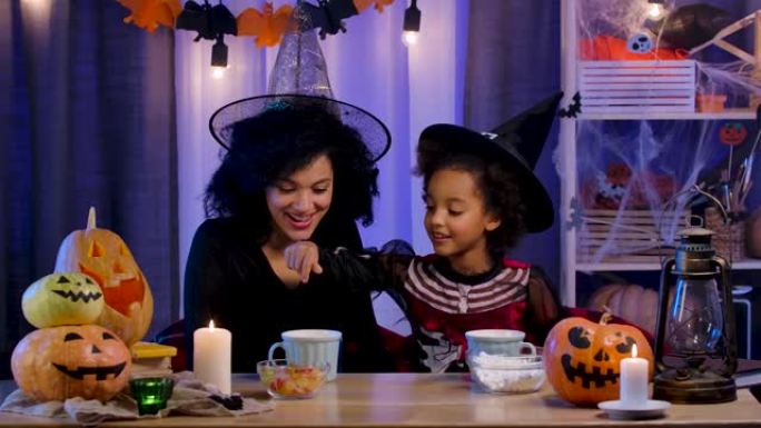 妈妈和女儿穿着节日服装和巫婆帽子的非洲裔美国人喝杯棉花糖的热饮。幸福的家庭坐在万圣节之夜装饰的房间的