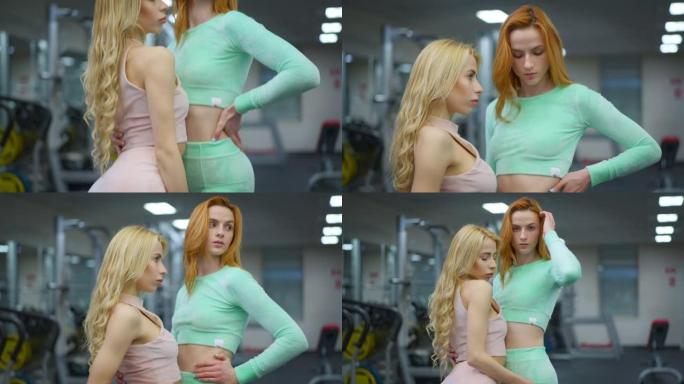 相机沿着两个漂亮自信的年轻白人女性在健身房摆姿势的苗条身材上升。自信健康的女运动员站在室内，面部表情