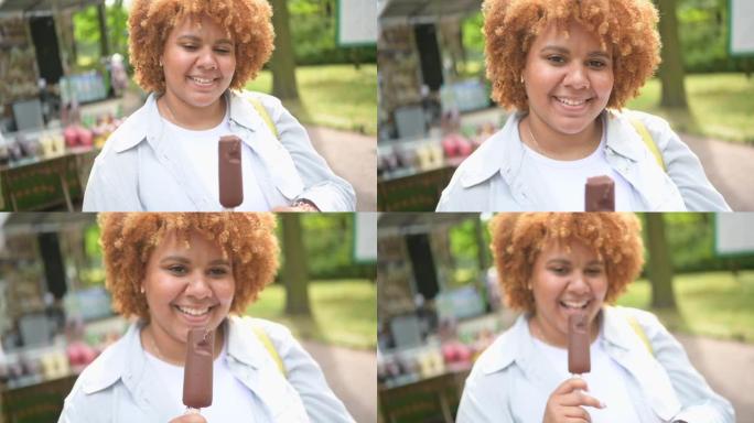 微笑的年轻自然美丽的又大的非洲裔美国妇女在公园吃冰淇淋巧克力冰棍的生活方式肖像。多样性和身体积极的概