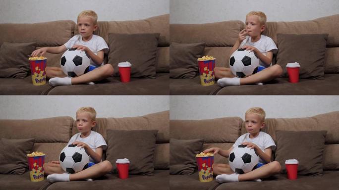 一个小男孩正坐在沙发上拿着爆米花在电视上看足球比赛。