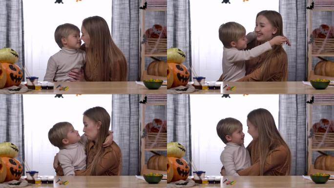 一名年轻女子坐在万圣节南瓜的桌子旁，将儿子抱在怀里。男孩亲吻他的母亲并紧紧拥抱她，她拥抱他。家庭爱情