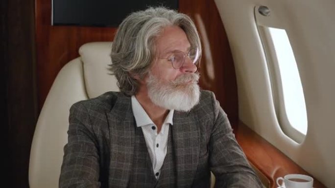 时尚成熟的百万富翁男子穿着古典西装豪华乘坐私人飞机飞行。自信的首席执行官商人透过公司喷气式飞机窗口。