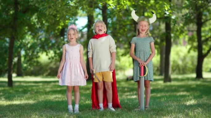 三个穿着服装的孩子看着相机，在公园里兴奋地尖叫。男孩戴着红色斗篷和国王王冠并手持剑，女孩戴着王冠和维