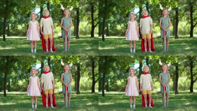 三个穿着服装的孩子看着相机，在公园里兴奋地尖叫。男孩戴着红色斗篷和国王王冠并手持剑，女孩戴着王冠和维