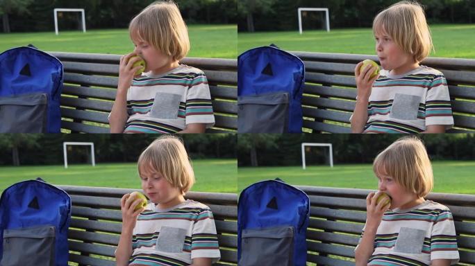 小男孩背着背包坐在学校院子附近的长凳上，放学后吃苹果作为零食
