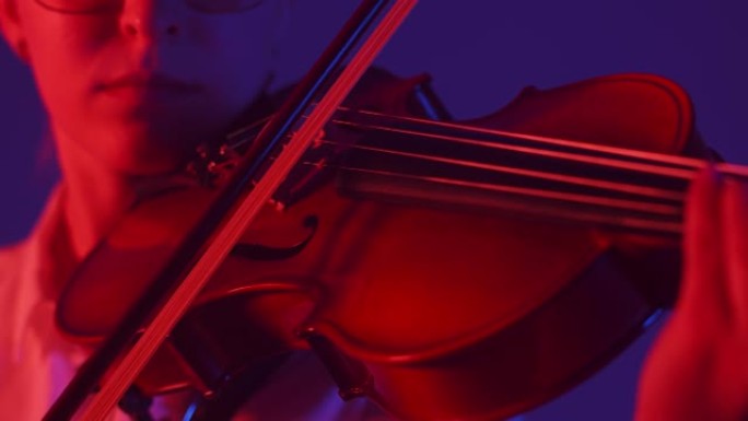 女音乐家在小提琴的琴弦上握着弓，在红灯的工作室里演奏交响音乐，特写镜头