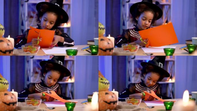 穿着节日服装和巫婆帽子的非洲裔美国小女孩用橙色纸切南瓜。孩子坐在万圣节之夜装饰的房间的桌子旁。特写。