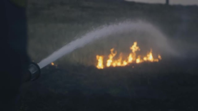 一名消防员手里拿着一根装有水的软管，扑灭了燃烧的火焰。自然物体正在燃烧: 草，草原，森林
