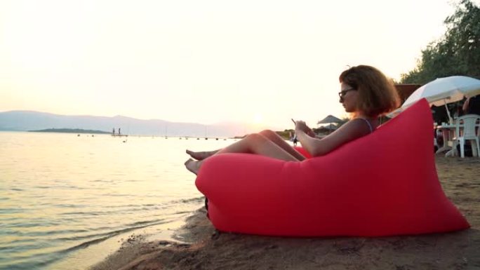 一个年轻女人在沙滩上悠闲地坐在红色躺椅上看书的4k视频片段充气太阳躺椅