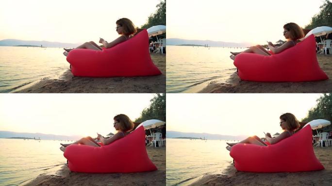 一个年轻女人在沙滩上悠闲地坐在红色躺椅上看书的4k视频片段充气太阳躺椅
