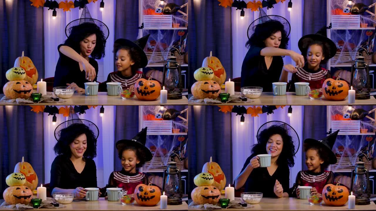 妈妈和女儿穿着节日服装和巫婆帽子的非洲裔美国人玩得开心，喝着杯子里的热饮。幸福的家庭坐在万圣节之夜装