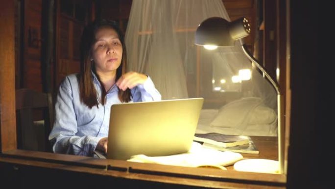 亚洲妇女洗澡后坐在窗户附近的桌子上，弄湿了黑发，继续工作到很晚，晚上用笔记本电脑做生意，感受积极的情