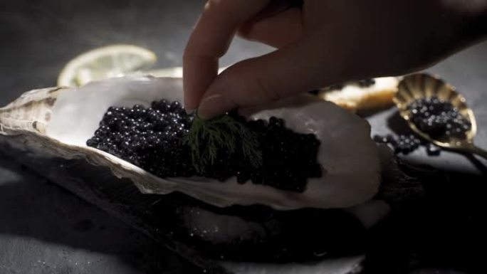 黑色鱼子酱在黑色背景的白色盘子上特写。一只雌性的手将一小枝莳萝放在黑色的鱼子酱上，鱼子酱站在黑色的木