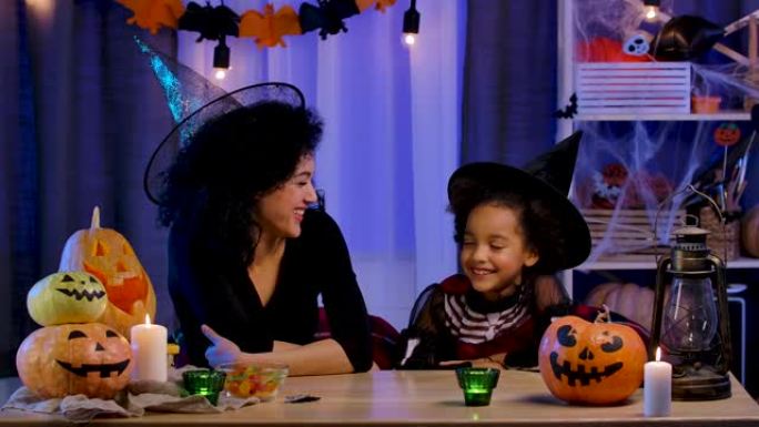 妈妈和女儿穿着节日服装和巫婆帽子的非洲裔美国人快乐地笑着玩得开心。幸福的家庭坐在万圣节之夜装饰的房间