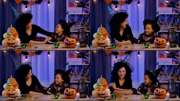 妈妈和女儿穿着节日服装和巫婆帽子的非洲裔美国人快乐地笑着玩得开心。幸福的家庭坐在万圣节之夜装饰的房间