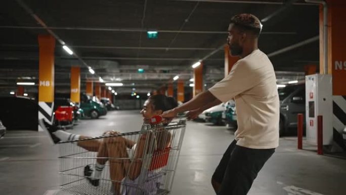 年轻的非洲裔美国人骑着他快乐的黑人女友在购物中心地下停车场的购物车里，慢动作