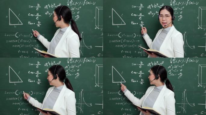 年轻的亚洲女教师拿着粉笔戴着耳机视频会议，配有学生摄像机。女教师培训课堂黑板在线课程中的数学。