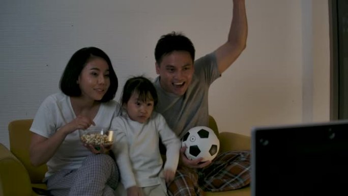亚洲家庭坐在沙发上观看电视足球比赛并一起加油，欢快地为胜利和胜利感到高兴。