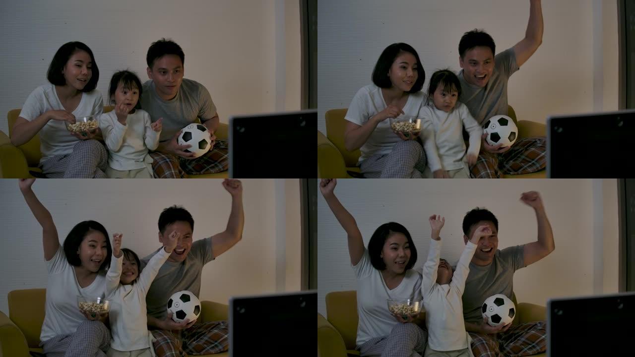 亚洲家庭坐在沙发上观看电视足球比赛并一起加油，欢快地为胜利和胜利感到高兴。