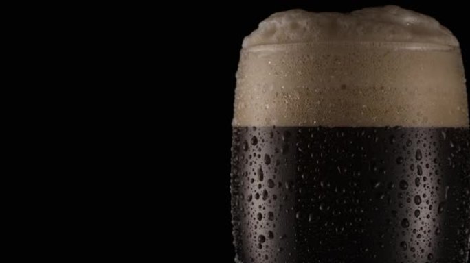 黑色背景上的一杯黑啤酒。啤酒在玻璃中摆动，气泡和泡沫上升。一杯啤酒顺时针缓慢旋转。