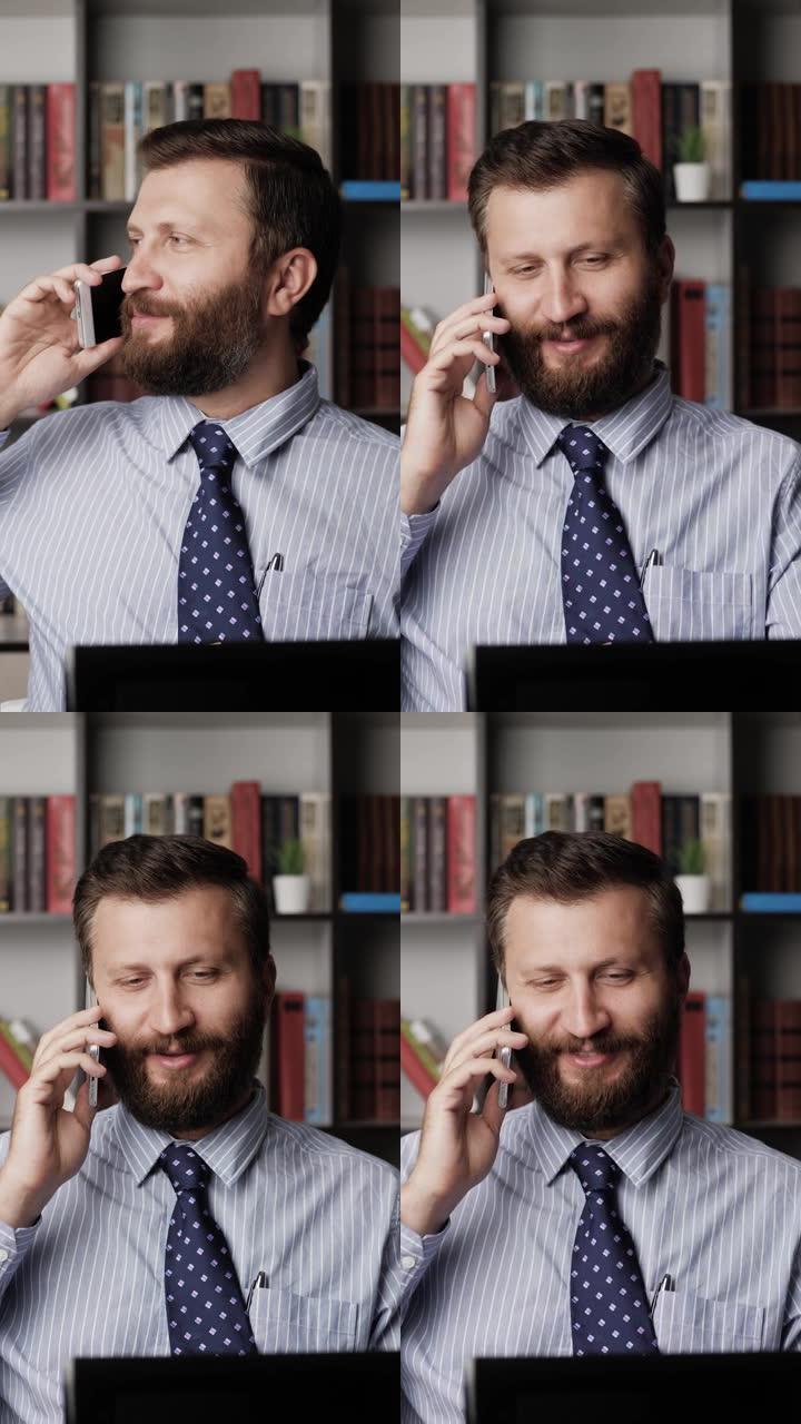 男人在打电话，微笑着。垂直查看工作场所积极的大胡子男人在电话上聊天，在笔记本电脑上打字。特写