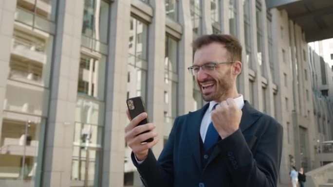 穿着正式西装的快乐兴奋的大胡子商人使用手机，用手显示是手势，从积极的情绪中惊呼，庆祝胜利，站在商业区
