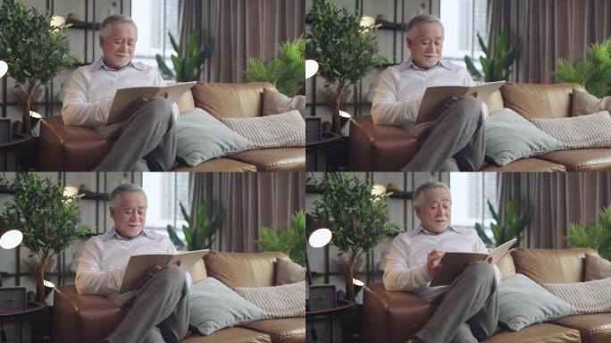 老亚洲高级祖父享受周日周末在家沙发沙发上阅读笔记本，在家隔离高级男性成人在家放松休闲快乐