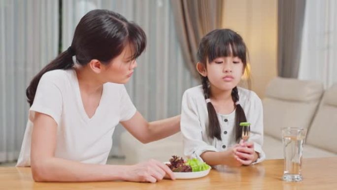 亚洲母亲教导和激励年幼的女孩在餐盘上吃绿色蔬菜。小女儿闻到黄瓜无聊的脸，摇头对妈妈说不。健康食品和儿