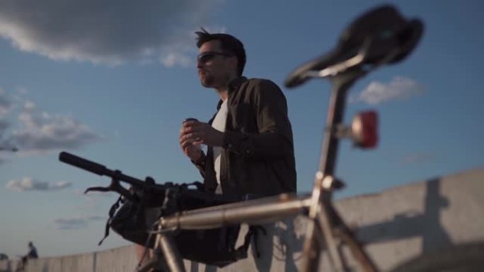 骑自行车的人坐在海边的水泥栅栏上，喝一次性咖啡，骑自行车时吃三明治。男子在湖边骑自行车后吃了午饭。下